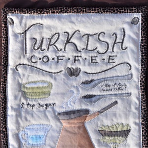 May include: A white fabric panel with a brown border, featuring a handwritten recipe for Turkish coffee. The recipe includes ingredients like ground coffee, sugar, water, and cardamom, illustrated with drawings of the ingredients and a coffee pot.