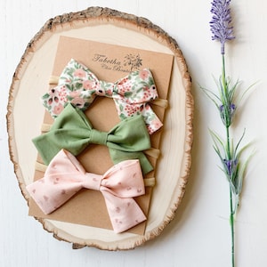 May include: Three baby headbands with bows in floral, solid green, and pink patterns. The headbands are displayed on a wooden slice with a sprig of lavender. The top headband has a floral pattern with pink and green flowers.