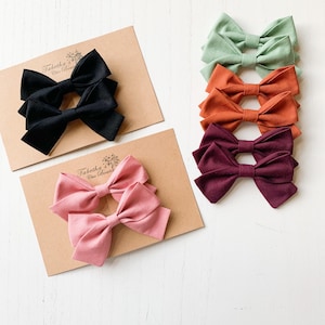 May include: Set of six fabric hair bows in various colors, including black, pink, burgundy, orange, and green. The bows are arranged on a white surface with brown cardstock behind them. The cardstock has the text "Tabitha Chic Boutique" printed on it.