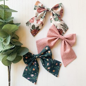 May include: Three fabric hair bows with floral patterns. The bows are in shades of pink, green, and brown. The bows are tied with a ribbon and are ready to be worn.