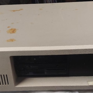 May include: A beige IBM desktop computer case featuring a rectangular chassis with a front-facing black drive bay, a ventilation grille on the left side, and a small IBM logo. The top surface shows several scattered brown rust-like stains.