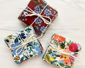 May include: Three bundles of fabric squares with floral patterns. Each bundle is tied with twine and features a different color scheme: red, blue, and green.
