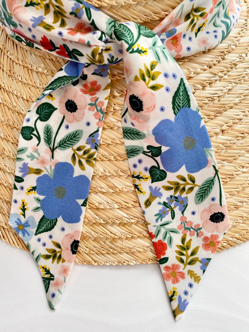 以下が含まれることがあります： 青、ピンク、緑の色合いの花柄が施された白い布のストール。 ストールは麦わら帽子にリボン結びされています。