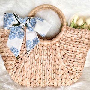 May include: Woven straw tote bag with a rounded handle. A white scarf with blue floral print is tied to the handle. The bag is filled with white flowers. The bag is sitting on a white, fluffy surface.