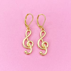 May include: A pair of gold-tone earrings featuring a treble clef design. The earrings have a simple, elegant style.
