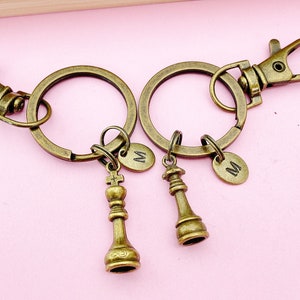 May include: Two bronze-colored keychains with chess piece charms. Each keychain has a large ring, a lobster clasp, and a small oval tag with the letter "M". One keychain features a king chess piece, and the other has a queen chess piece.