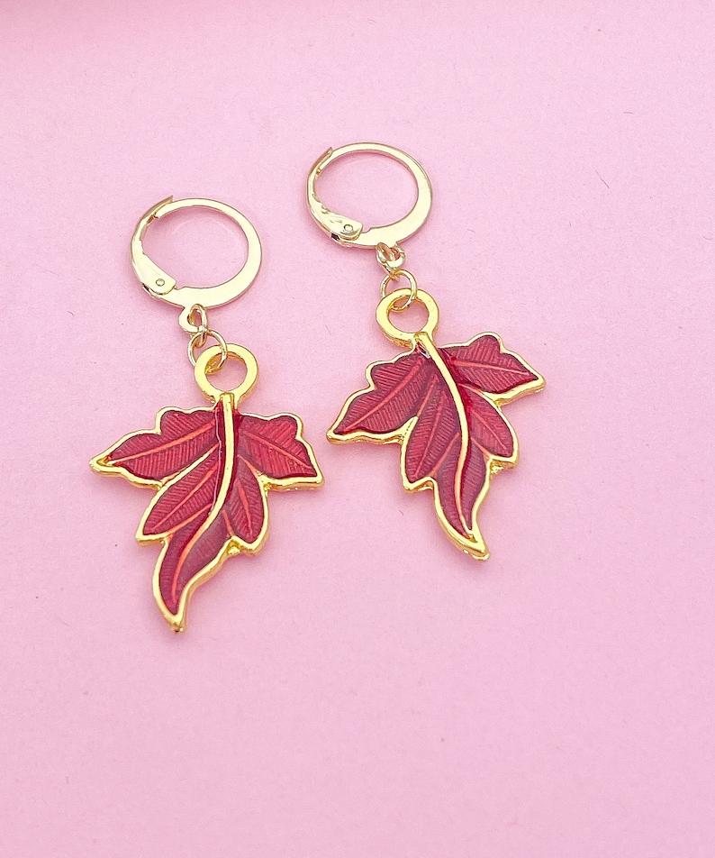 May include: A pair of gold earrings with red leaf designs. The leaves are detailed with gold veins and have a glossy finish.