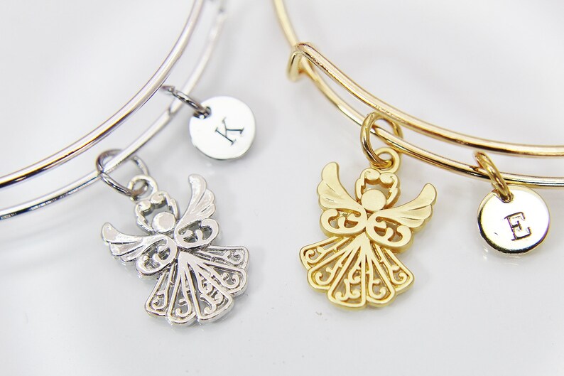 K&ouml;nnte beinhalten: Zwei dehnbare Armreifen mit Engelsanh&auml;ngern und personalisierten Buchstaben-Scheiben. Das Armband links ist silberfarben mit einem silbernen Engelsanh&auml;nger und einer Scheibe mit dem Buchstaben "K". Das Armband rechts ist golden mit einem goldenen Engelsanh&auml;nger und einer Scheibe mit dem Buchstaben "E".