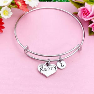 May include: A silver-toned adjustable bangle bracelet with two charms. One charm is a heart with the word "Nanny" and a small clear gem. The other charm is a small round disc with the letter "L".