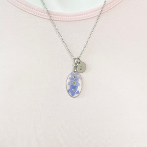 May include: A silver chain necklace with a silver oval pendant containing pressed forget-me-not flowers and a silver initial charm with the letter "J".