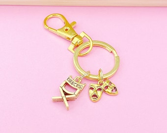 May include: Gold-colored keychain with a lobster clasp, key ring, and three charms. Charms include a director's chair with the word "DIRECTOR", and two theater masks. The background is a light pink color.