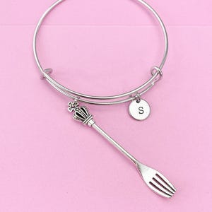 May include: Silver charm bracelet with a fork charm and a crown charm. The charm bracelet has a round charm with the letter "S" on it.