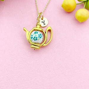 May include: A gold-colored teapot pendant necklace with a gold chain. The teapot holds a white and blue floral design. A small gold charm with the letter "Y" hangs from the chain. The background is pink with yellow lemons and pink flowers.