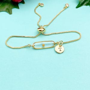 May include: A gold-tone adjustable slider bracelet featuring a thin box chain, a clear rectangular resin charm containing a single small yellow bead, and a small round gold-tone disc charm engraved with the capital letter T.