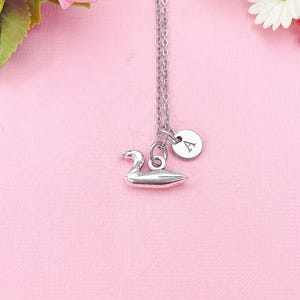 May include: A silver-colored necklace with a small swan pendant and a round charm with the letter "A". The necklace hangs against a pink background, with pink roses and white flowers in the background.