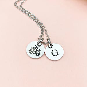 May include: A silver-colored necklace with two round pendants. One pendant features a drum set graphic, and the other has the letter "G" in a bold font. The chain is delicate, and the pendants are connected. The background is a soft pink.