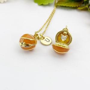 May include: A gold-colored necklace featuring two pendants. One pendant is a round, orange-colored locket with gold accents. The other pendant is a round charm with the letter "U". The second pendant is an open locket with a heart-shaped gem.