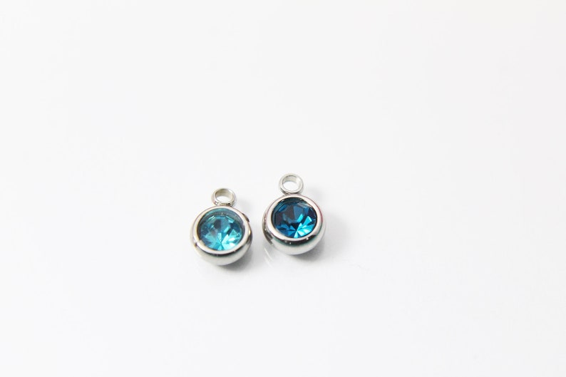 May include: Two silver charms with a blue gemstone in the center. The charms have a loop at the top for attaching to a bracelet or necklace.