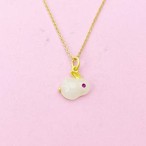 May include: A gold chain necklace with a white rabbit pendant. The rabbit has a pink gemstone for an eye.