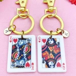 May include: Two keychains with gold-colored hardware. Each keychain has a playing card design: a King and a Queen of Hearts. Each keychain has a small, round charm with a letter. The background is a light pink color.