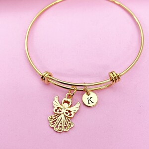 K&ouml;nnte beinhalten: Goldenes Armreif-Armband mit einem Engelsanh&auml;nger und einem kleinen runden Anh&auml;nger mit dem Buchstaben K.