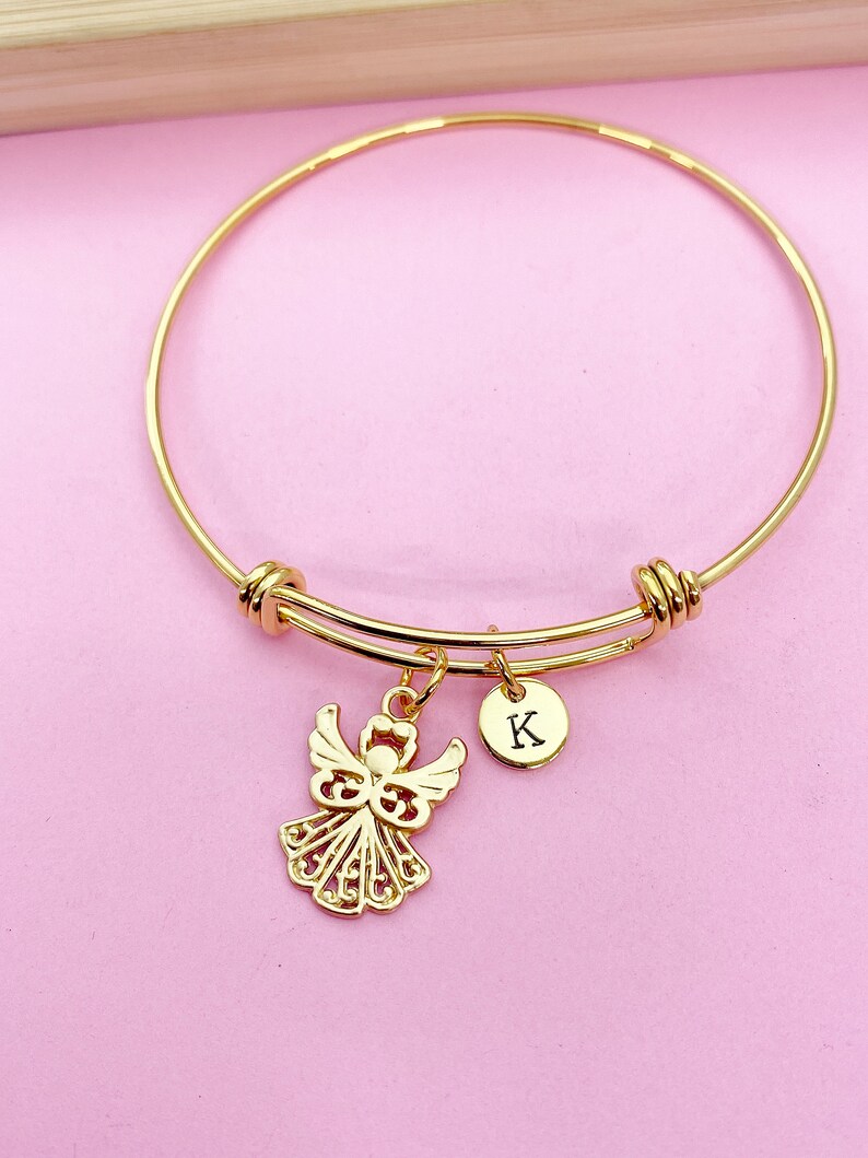 K&ouml;nnte beinhalten: Goldenes Armreif-Armband mit einem Engelsanh&auml;nger und einem kleinen runden Anh&auml;nger mit dem Buchstaben K.