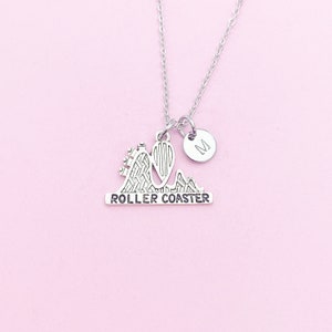 May include: Silver-toned necklace with a roller coaster pendant and a small, round charm with the letter "M". The roller coaster pendant has the word "ROLLER COASTER" engraved on it. The necklace is displayed on a light pink background.