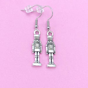 May include: A pair of silver-toned nutcracker earrings with a hook closure. The earrings are on a pink background.