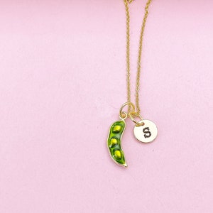 Peut inclure: Un collier dor&eacute; avec un pendentif en forme de cosse de pois verte contenant trois petits pois jaunes, et un petit charme rond avec la lettre "S". Le collier est pr&eacute;sent&eacute; sur un fond rose.