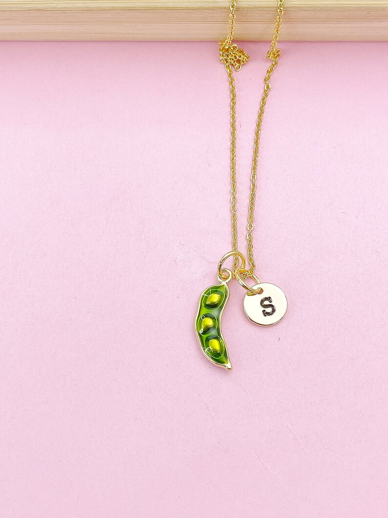 Peut inclure: Un collier dor&eacute; avec un pendentif en forme de cosse de pois verte contenant trois petits pois jaunes, et un petit charme rond avec la lettre "S". Le collier est pr&eacute;sent&eacute; sur un fond rose.