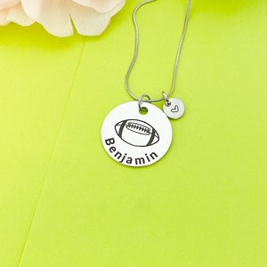 May include: A silver necklace with a round pendant featuring a football design and the name "Benjamin" engraved on it. A small heart charm hangs from the pendant.