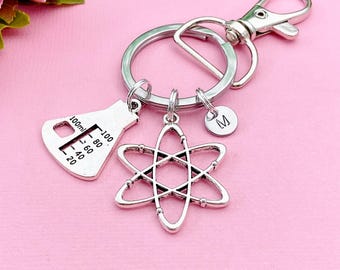 May include: Silver-colored science-themed keychain with three charms: a flask with measurements, an atom symbol, and a small disc with the letter "M". The keychain has a clasp and a split ring.
