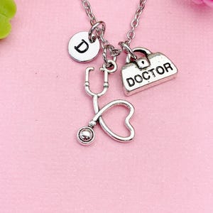 May include: Silver-colored necklace with a stethoscope charm, a charm with the word "DOCTOR", and a round charm with the letter "D". The necklace is displayed on a pink background, with a pink rose in the background.