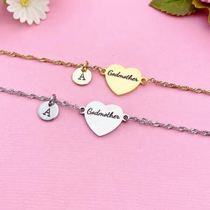 May include: Two delicate bracelets with heart-shaped pendants that read "Godmother." One is gold-toned, the other silver-toned. Each bracelet has a small, round charm with the letter "A." The background is a soft pink.