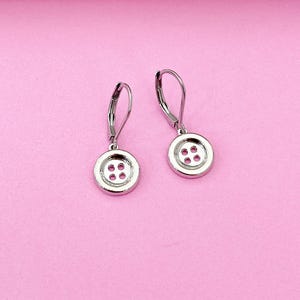 May include: A pair of silver button-shaped earrings with four holes in each button. The earrings are on a pink background.