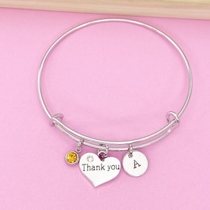 May include: Silver bangle bracelet with a heart charm that says "Thank you", a small round charm with a single letter "A", and a small yellow gemstone charm.
