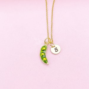 Peut inclure: Un collier dor&eacute; avec un pendentif en forme de cosse de pois verte et un charm circulaire avec la lettre "S". La cosse de pois contient trois petits pois. Le fond est rose clair.