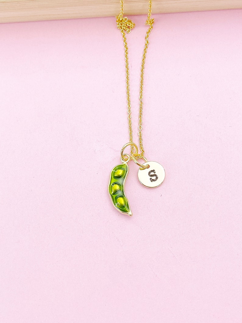 Peut inclure: Un collier dor&eacute; avec un pendentif en forme de cosse de pois verte et un charm circulaire avec la lettre "S". La cosse de pois contient trois petits pois. Le fond est rose clair.
