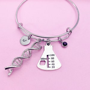 May include: Silver-toned adjustable bangle bracelet with science-themed charms. Charms include a DNA helix, a flask with 100ml markings, a small initial disc with the letter "J", and a dark purple gemstone. The bracelet is displayed on a pink background.