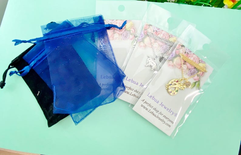 May include: Two blue organza bags, one black velvet bag, and two clear plastic bags with jewelry inside. The jewelry is a silver necklace with a charm and a gold necklace with a charm. The plastic bags have a white card with the text "Lebue Jewelry" and the website address "www.lebuejewelry.com".