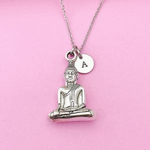 May include: Silver necklace with a small Buddha pendant and a charm with the letter A. The necklace is on a pink background.