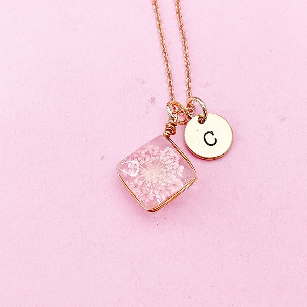May include: A rose gold necklace with a pendant featuring a clear, square resin encasing delicate white and pink flowers. A small, round charm with the letter "C" hangs next to the square pendant. The necklace is displayed on a pink background.