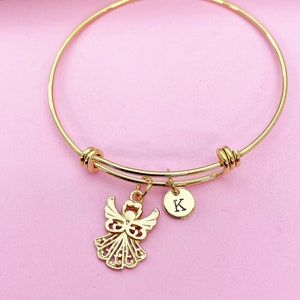 K&ouml;nnte beinhalten: Goldenes Armreif-Armband mit einem Engelsanh&auml;nger und einem kleinen runden Anh&auml;nger mit dem eingravierten Buchstaben "K".