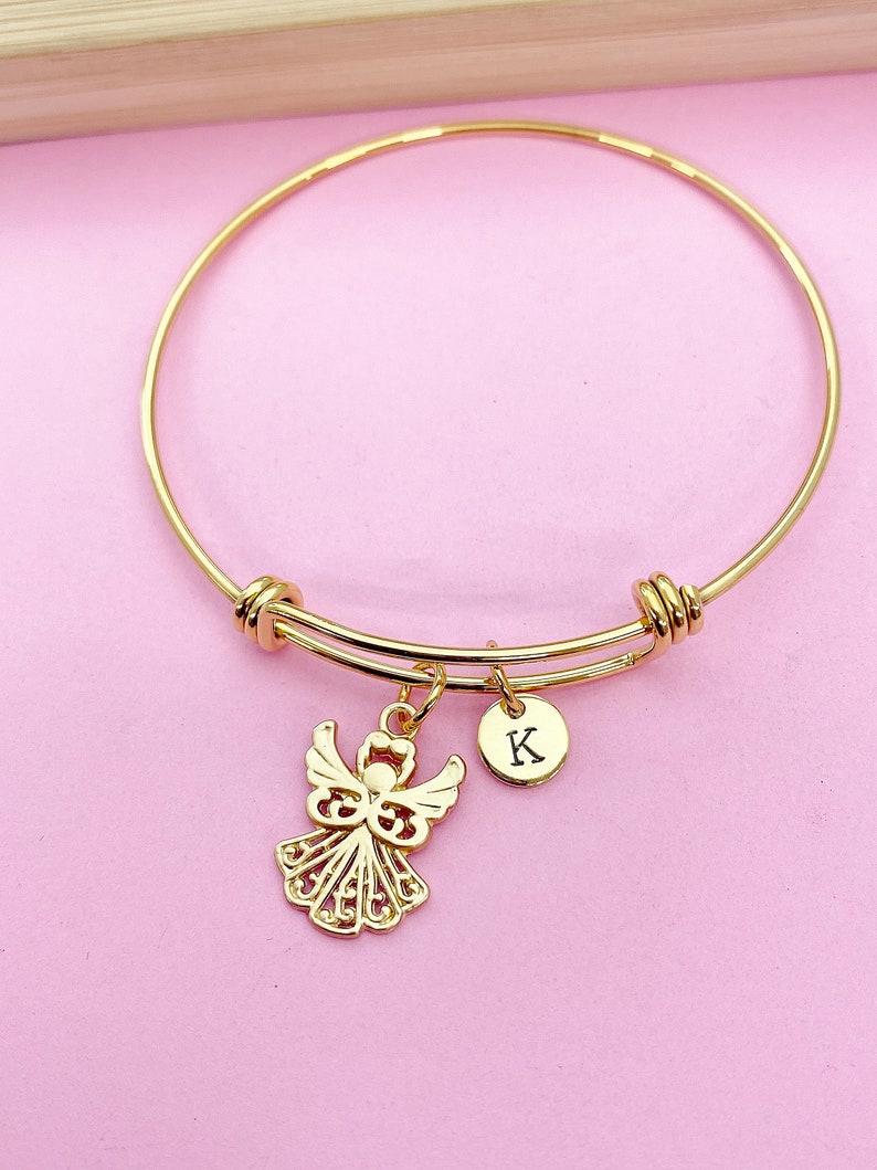 K&ouml;nnte beinhalten: Goldenes Armreif-Armband mit einem Engelsanh&auml;nger und einem kleinen runden Anh&auml;nger mit dem eingravierten Buchstaben "K".