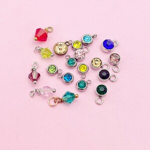 May include: Assortment of colorful crystal charms with silver jump rings. The charms feature various shapes and sizes of crystals, including round, square, and teardrop.