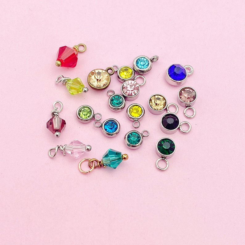 May include: Assortment of colorful crystal charms with silver jump rings. The charms feature various shapes and sizes of crystals, including round, square, and teardrop.