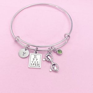 May include: Silver-toned bangle bracelet with multiple charms. Charms include a round initial charm with the letter "Y", a miniature eye chart, a pair of eyeglasses, and a small green gemstone. The bracelet is displayed on a pink background.