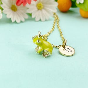 May include: A gold-colored necklace featuring a small, bright green frog pendant. The frog has detailed legs and a textured body. A circular charm with the letter "U" is also attached. The background includes white flowers and small oranges.