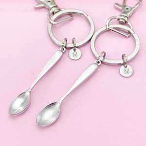 May include: Two silver spoon keychains with silver key rings and silver clasps. Each spoon has a small silver charm with the letter "M" on it.