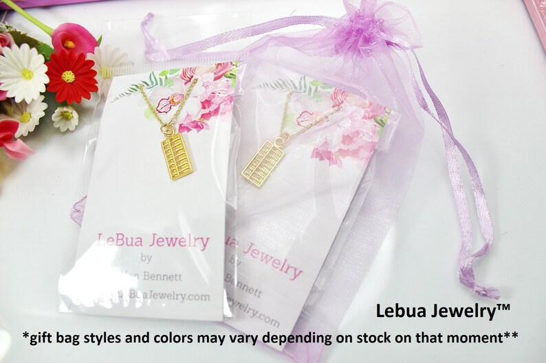 K&ouml;nnte beinhalten: Zwei goldene Abakus-Halsketten in durchsichtiger Plastikverpackung mit rosafarbenem Blumendesign. Die Halsketten sind in einem lila Organzas&auml;ckchen verpackt. Die Verpackung enth&auml;lt den Text "LeBua Jewelry by Bennett Bua.Jewelry.com" und "Lebua Jewelry&trade; *Geschenkbeutel-Stile und -Farben k&ouml;nnen je nach Lagerbestand variieren**"
